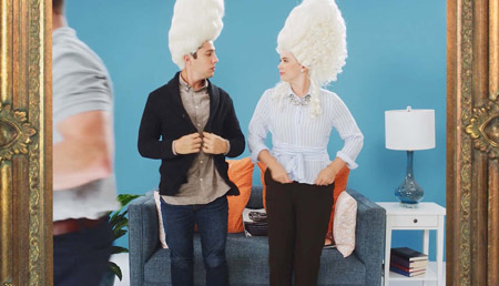 A still of a dolly shot pulling away from a couple wearing modern clothes and ornate 18th century style wigs. Behind them is a set decorated to look like a living room with a couch and decor. A crew member walks in front of them. They're surrounded by a frame